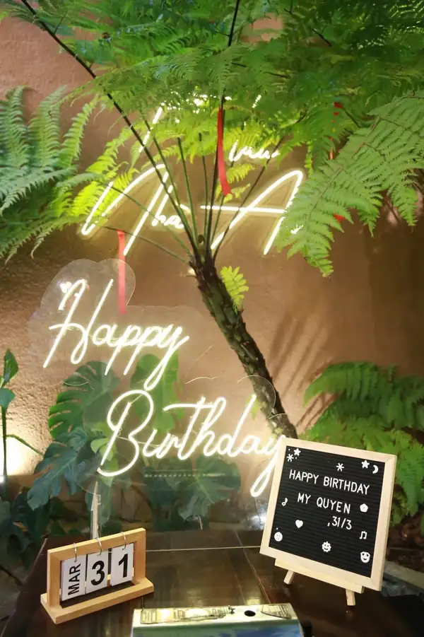 Đèn neon Nhà An và Happy Birthday sáng trên nền cây xanh, bên dưới là bảng viết tên khách